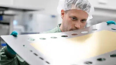 A man wearing protective clothing is holding a bipolar plate in a factory producing electrolysis components.